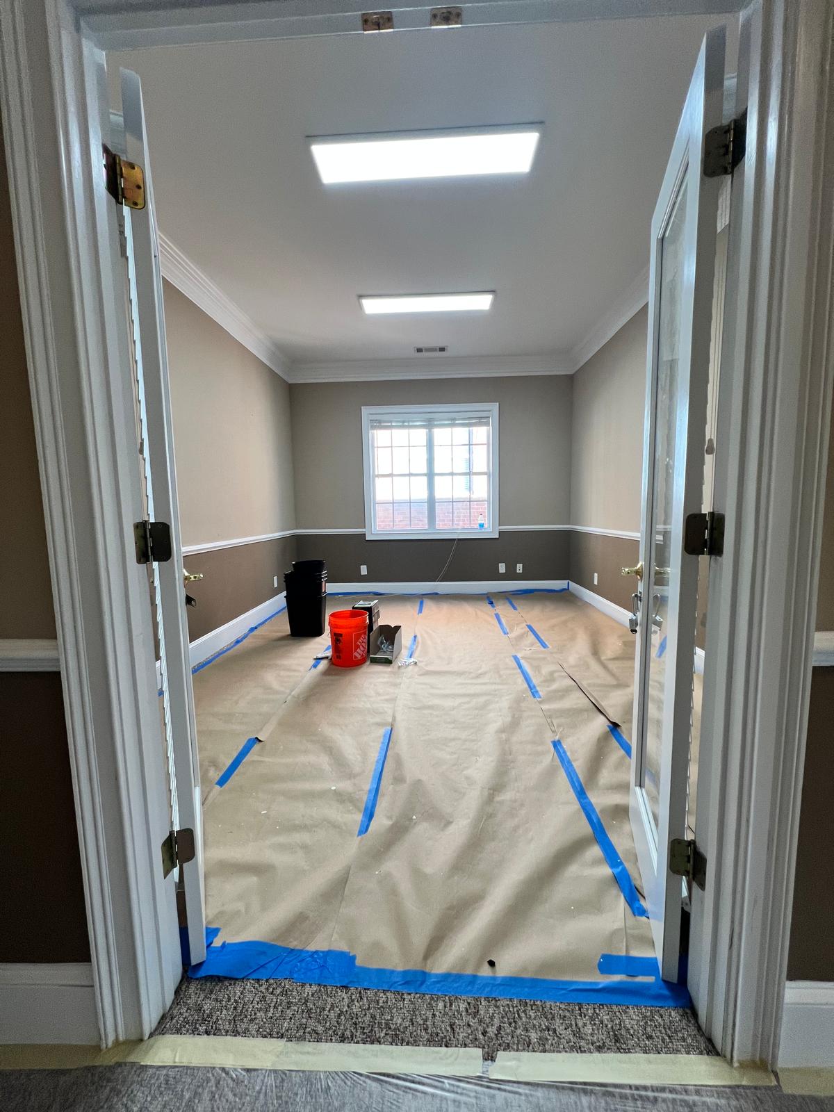 Office room viewed through double doors with floor protection paper, blue tape and painting materials in place