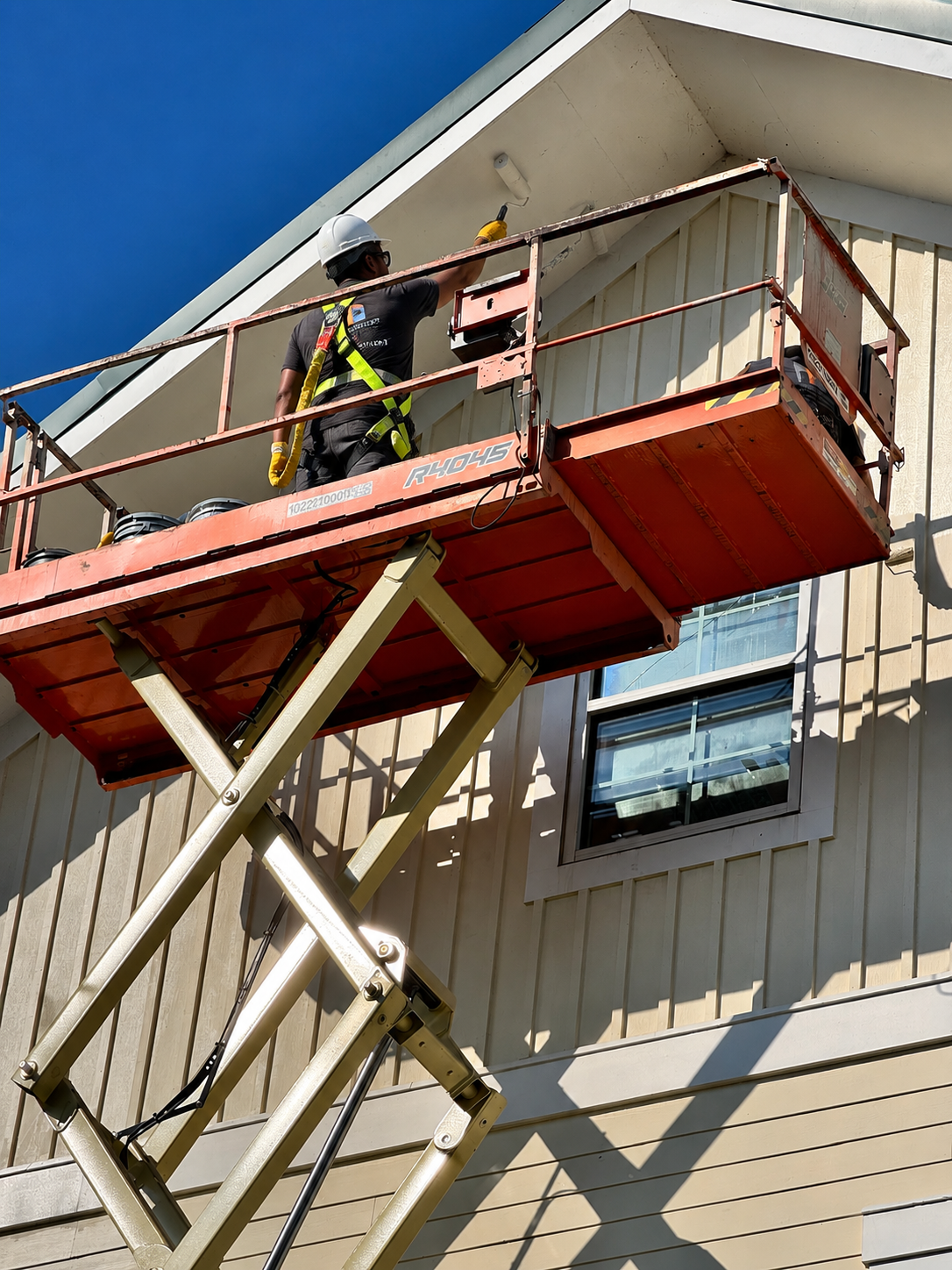 Close-up of a DJ7 Solutions worker in a scissor lift basket painting exterior trim and siding under a gable