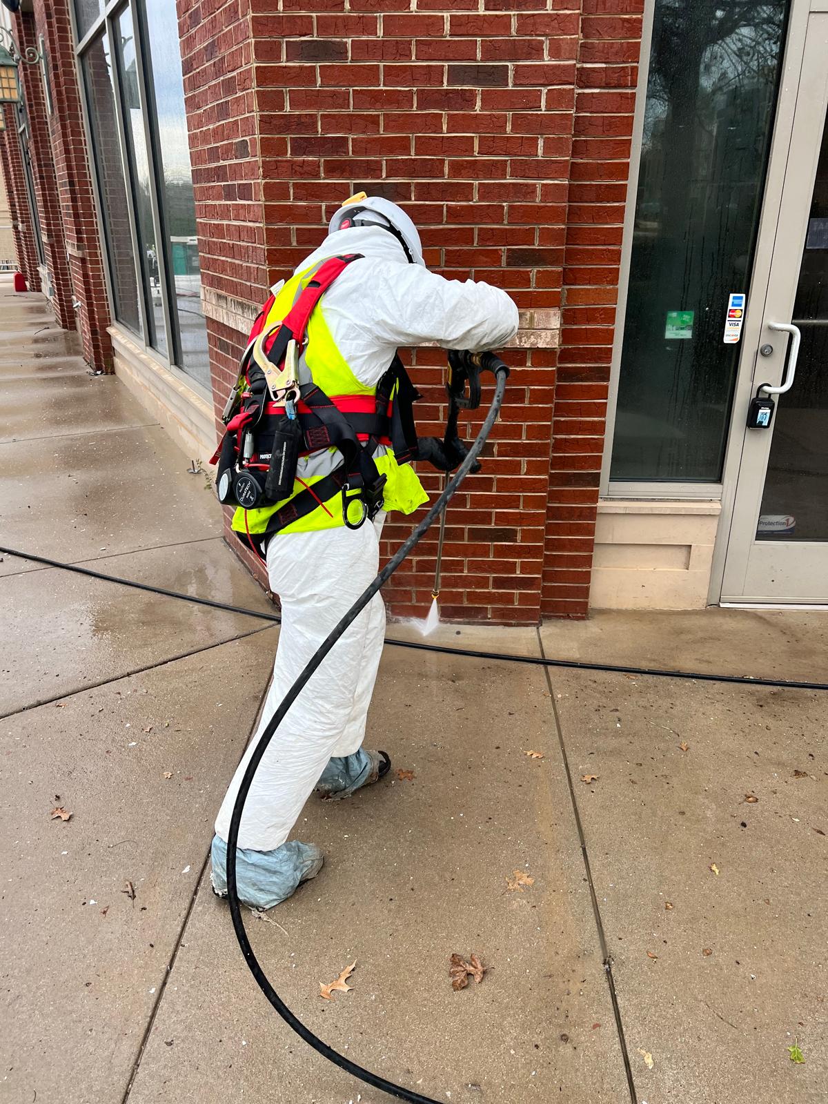 DJ7 Solutions technician in protective gear pressure washing a brick building exterior and ground-level walkway