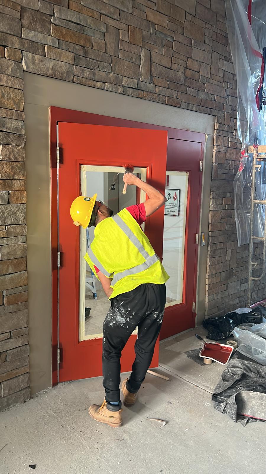 Worker painting a red commercial exterior door set in a stone wall with drop cloths in place